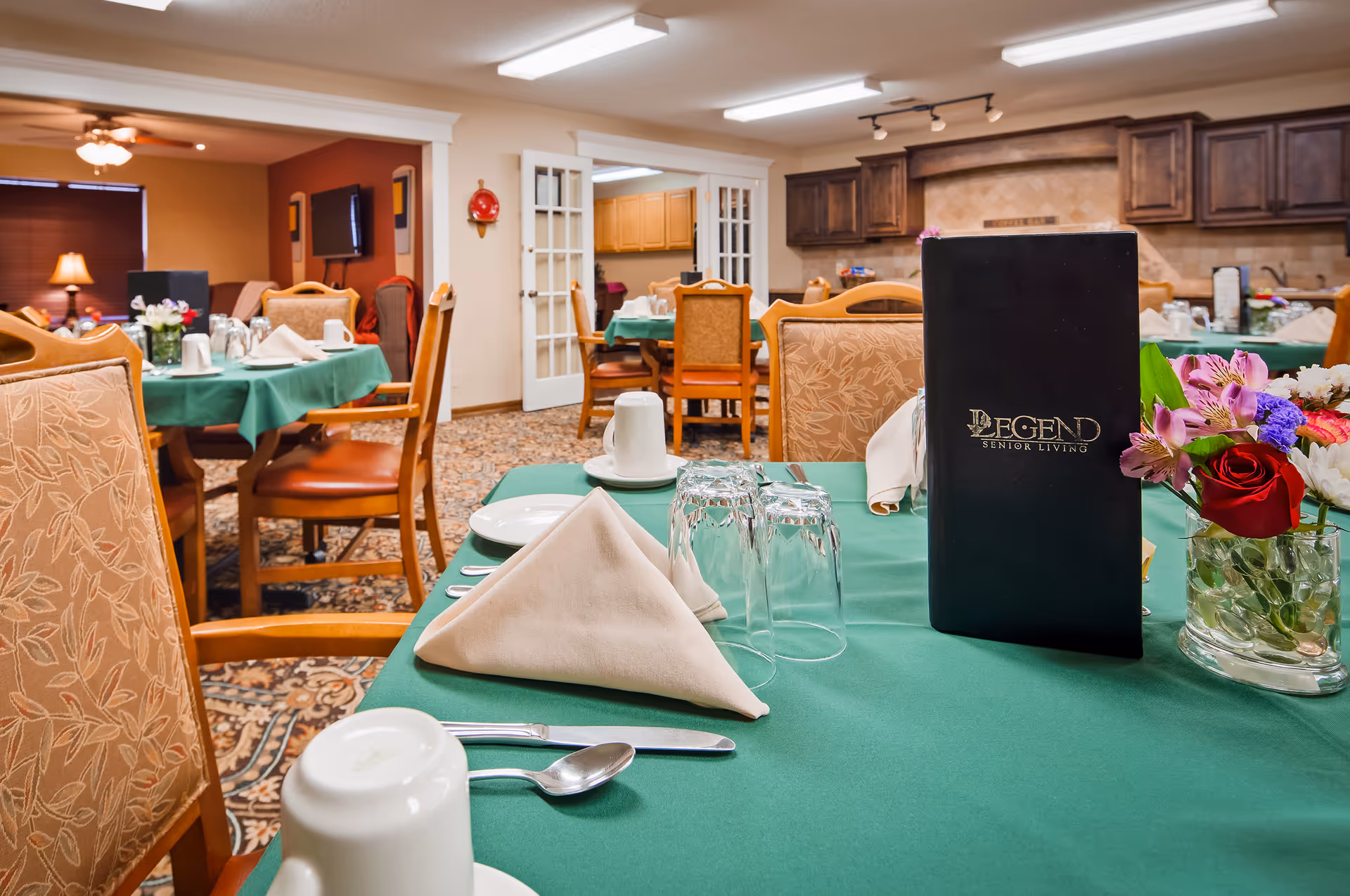 Dining room with tables covered in green tablecloths, place settings, floral centerpieces, and a visible 'Legend Senior Living' menu.
