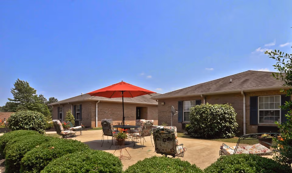 Outdoor patio area at Dogwood Bend with cushioned chairs and a table under a red umbrella, surrounded by green bushes and two single-story brick buildings under a clear blue sky.