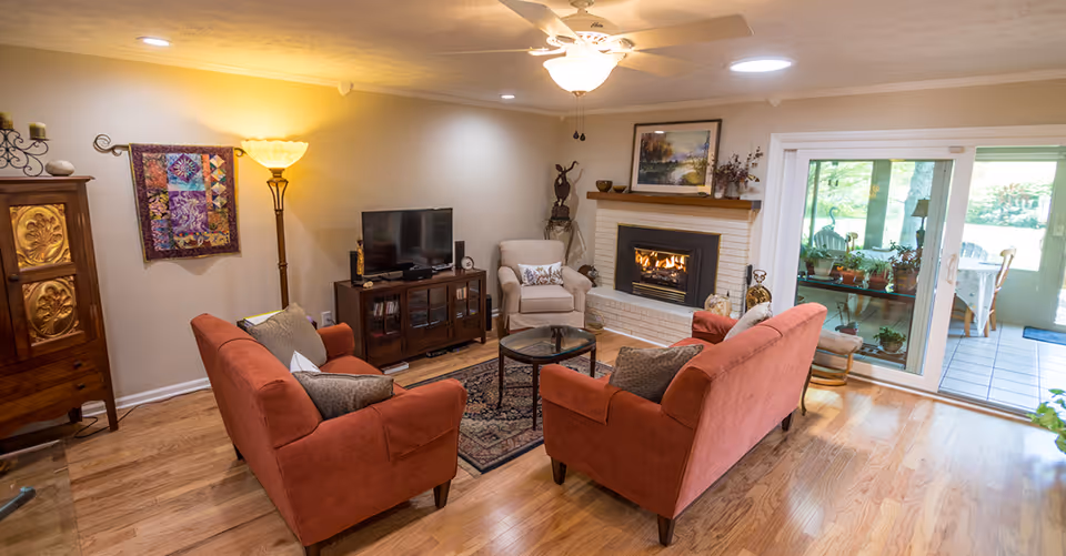 A cozy living room with two red sofas and a beige armchair arranged around a glass coffee table on a patterned rug. There is a fireplace with a fire burning, a TV on a wooden stand, a floor lamp, and a wooden cabinet. Sliding glass doors lead to a sunroom with plants and outdoor furniture visible.