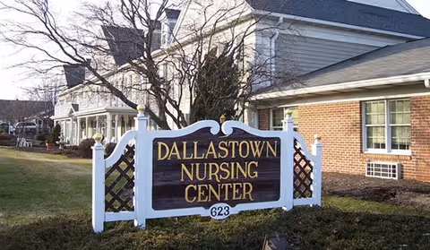 Outdoor view of the Dallastown Nursing Center sign in front of a building with white siding and brick exterior walls, with leafless trees and grass around.