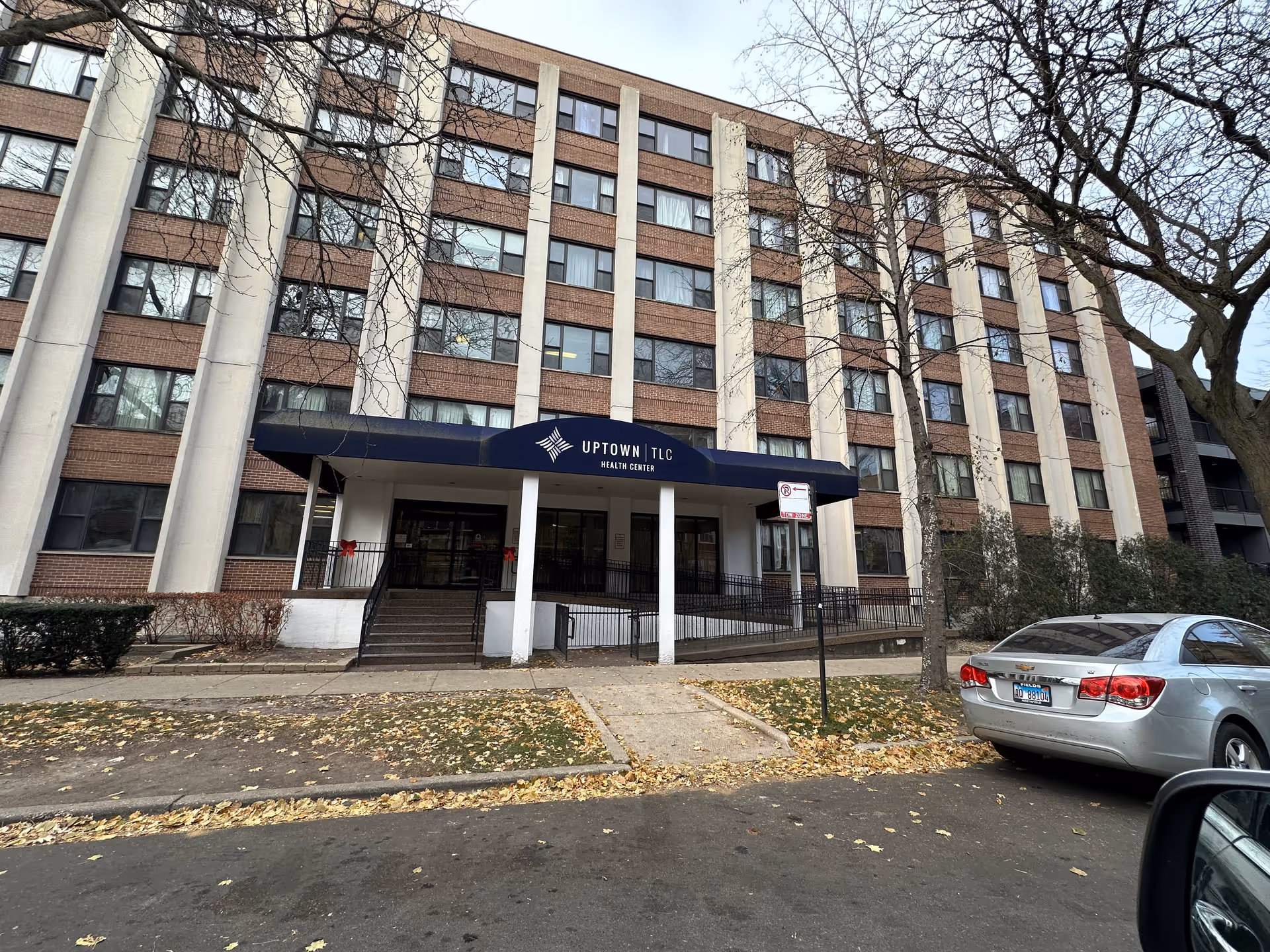 Front exterior of a multi-story brick care and rehab building with a blue entrance awning that reads 'Uptown TLC Health Center' and a car parked on the street.