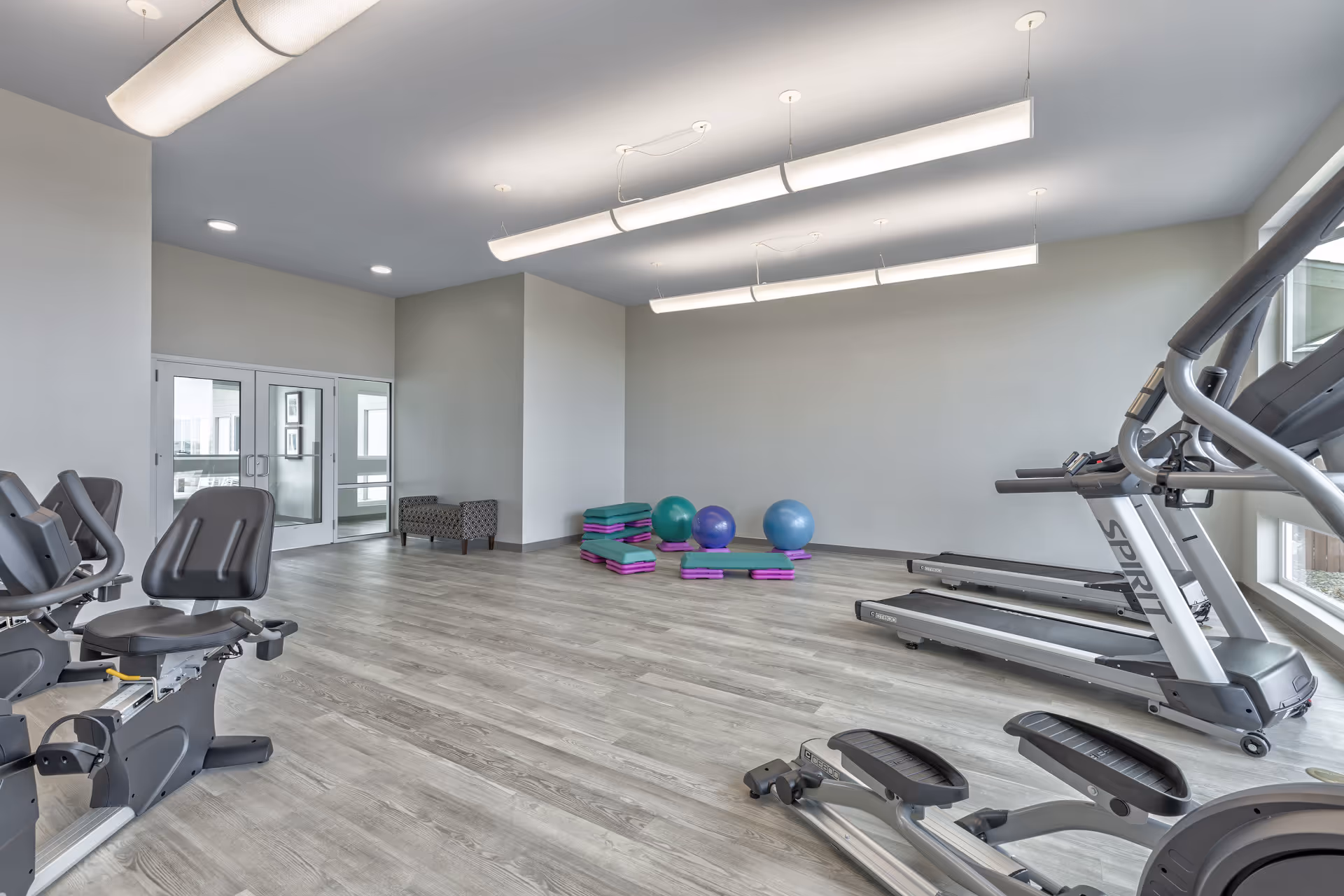 A spacious fitness room with light wood flooring and white walls. The room contains exercise equipment including treadmills, a recumbent bike, and an elliptical machine. In the back corner, there are stacked aerobic steps and three exercise balls in blue and purple. Large windows on the right side allow natural light to enter the room. Modern ceiling lights hang from the ceiling, and there is a double glass door leading to another area.
