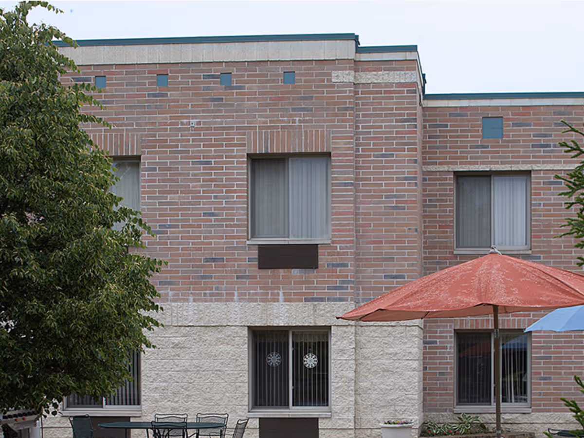 Exterior view of a brick building with two windows on the upper floor and two windows on the lower floor. There is a large tree on the left side and two patio umbrellas, one red and one blue, shading outdoor tables and chairs in front of the building.