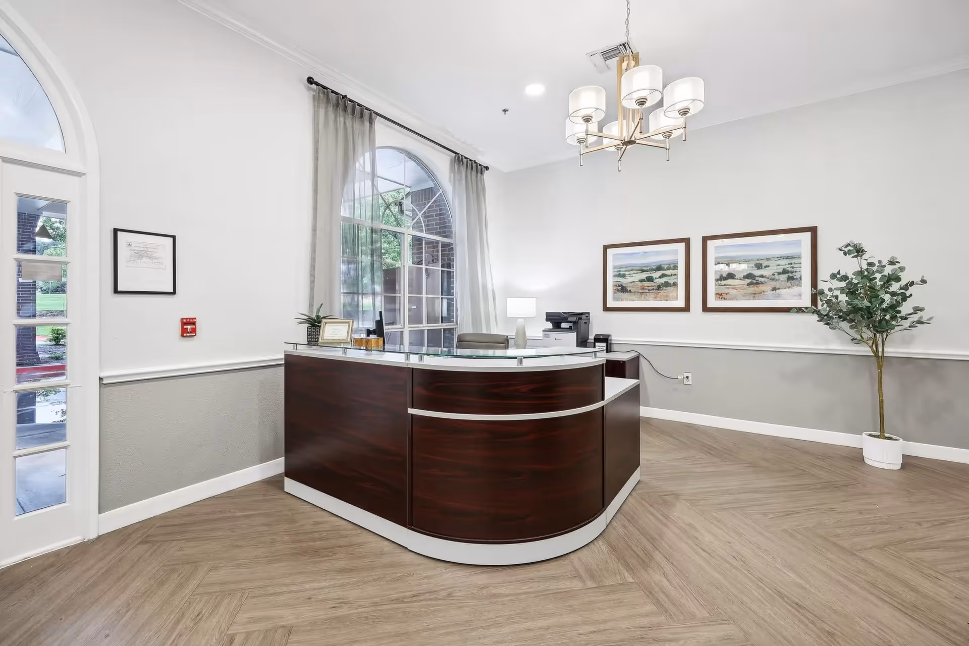 Reception area with a curved dark wood desk, a lamp, a printer, and framed landscape pictures on the wall. There is a large arched window with sheer curtains and a potted plant in the corner. The floor has a light wood pattern and a modern chandelier hangs from the ceiling.