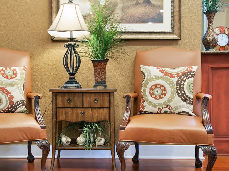 Two brown leather armchairs with decorative patterned pillows flank a wooden side table. On the table is a lamp with a beige shade and a potted green plant. Behind the table is a framed painting on a beige wall. To the right, part of a wooden cabinet with a decorative clock and vase is visible.