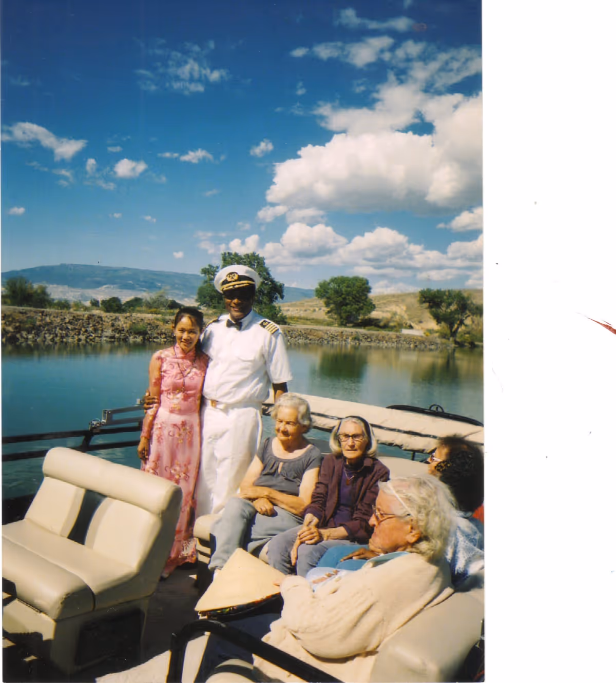 A group of elderly people sitting on a boat with a man in a captain's uniform and a woman in a pink dress standing beside them. The boat is on a calm body of water with trees and hills in the background under a partly cloudy blue sky.