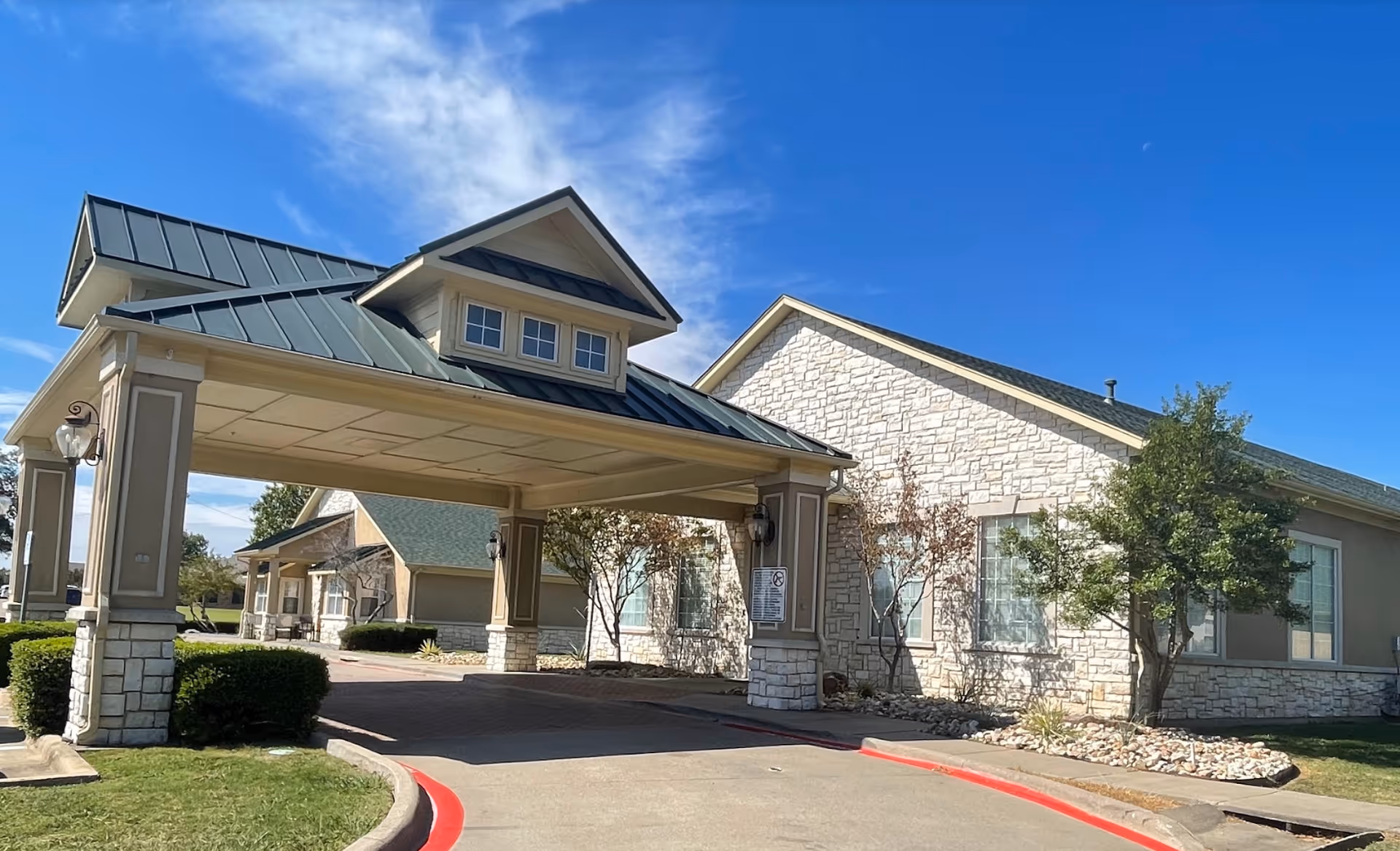 Exterior view of Colonial Lodge Assisted Living facility showing a covered driveway entrance with a peaked roof, beige stone and siding walls, windows, small trees, and a clear blue sky.
