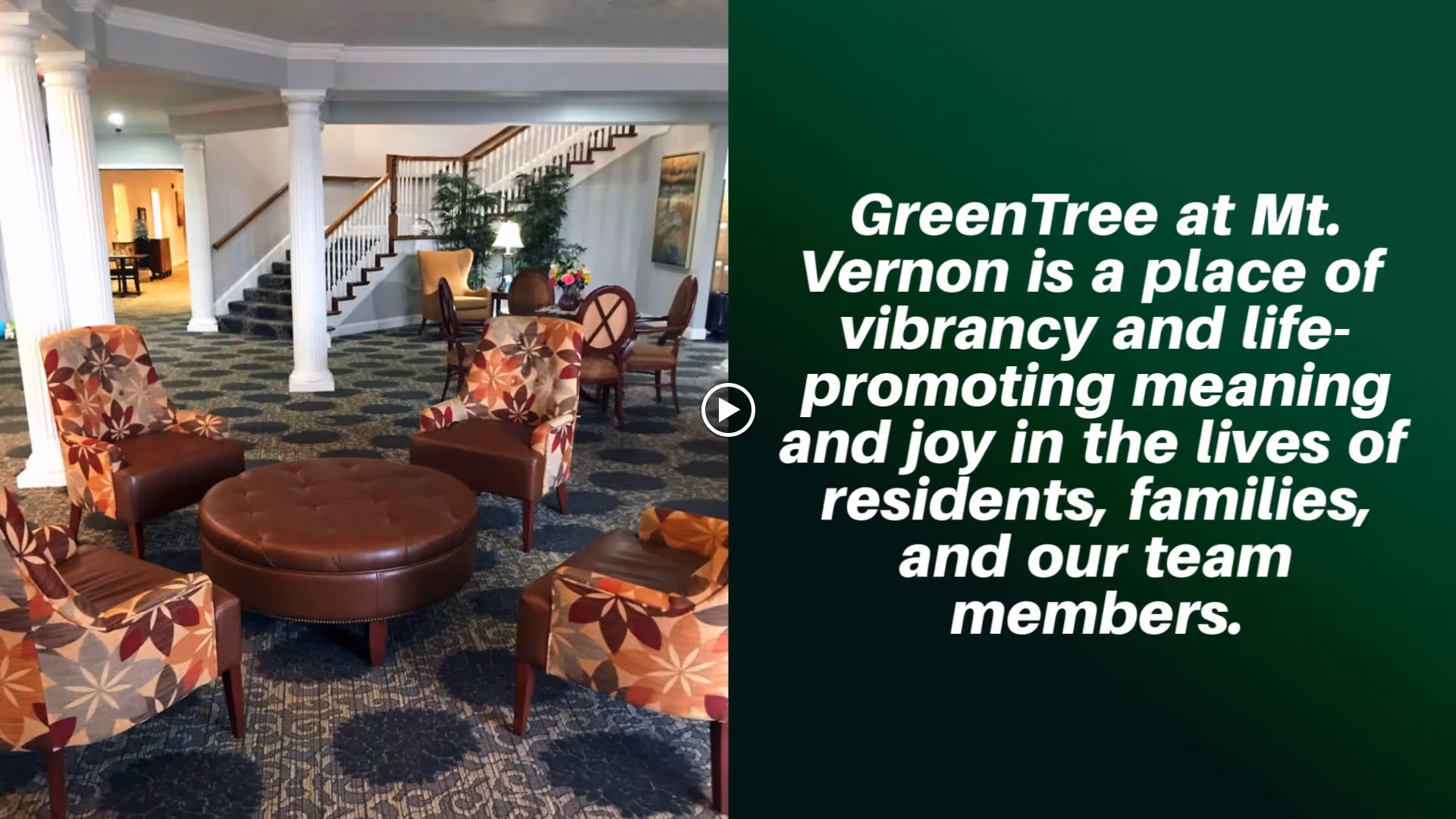 A spacious and well-lit common area in a senior living facility featuring four patterned armchairs arranged around a large round brown ottoman. In the background, there is a staircase with wooden railings, a table with chairs, potted plants, and framed artwork on the wall.