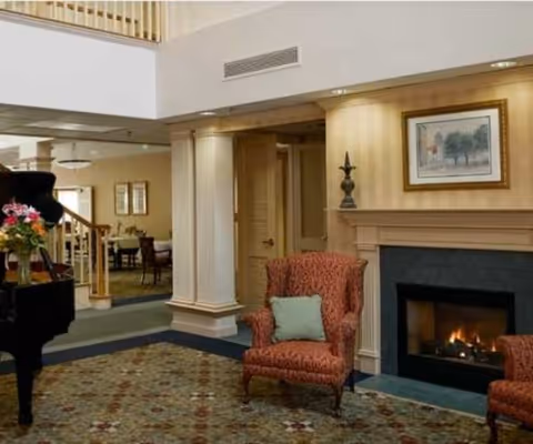A cozy living room area with a lit fireplace, two red patterned armchairs with light blue cushions, a decorative lamp on the mantel, and a framed painting above the fireplace. In the background, there is a dining area with tables and chairs, and a grand piano with a vase of flowers on it.