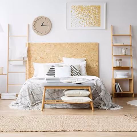 A bright and minimalist bedroom featuring a bed with a wooden headboard, white and patterned pillows, and a gray blanket. There is a wooden bench at the foot of the bed with two pillows underneath. On either side of the bed are wooden ladder-style shelves holding various decorative items. A round beige clock and a framed abstract gold and white artwork hang on the white wall above the bed. The floor has a woven beige rug.