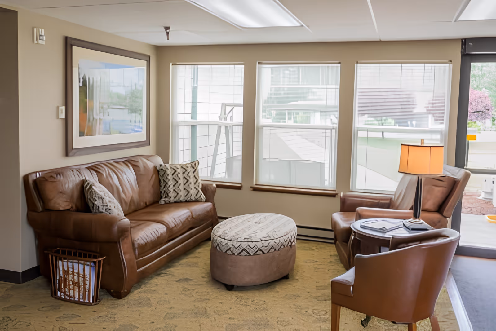 A cozy living room area with a brown leather sofa adorned with patterned pillows, a matching armchair, and a round ottoman in the center. A small round side table with a lamp and some magazines is next to the armchair. Three large windows with white blinds allow natural light to fill the room, and a framed picture hangs on the wall above the sofa.