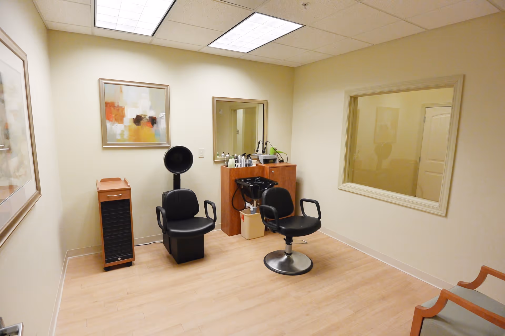 A small hair salon room with two black salon chairs, a hair washing station with a sink, a wooden cabinet with hair care products, and a large mirror on the wall. The room has light-colored walls, a wooden floor, and two framed abstract paintings. There is a large window on one wall looking into another room.