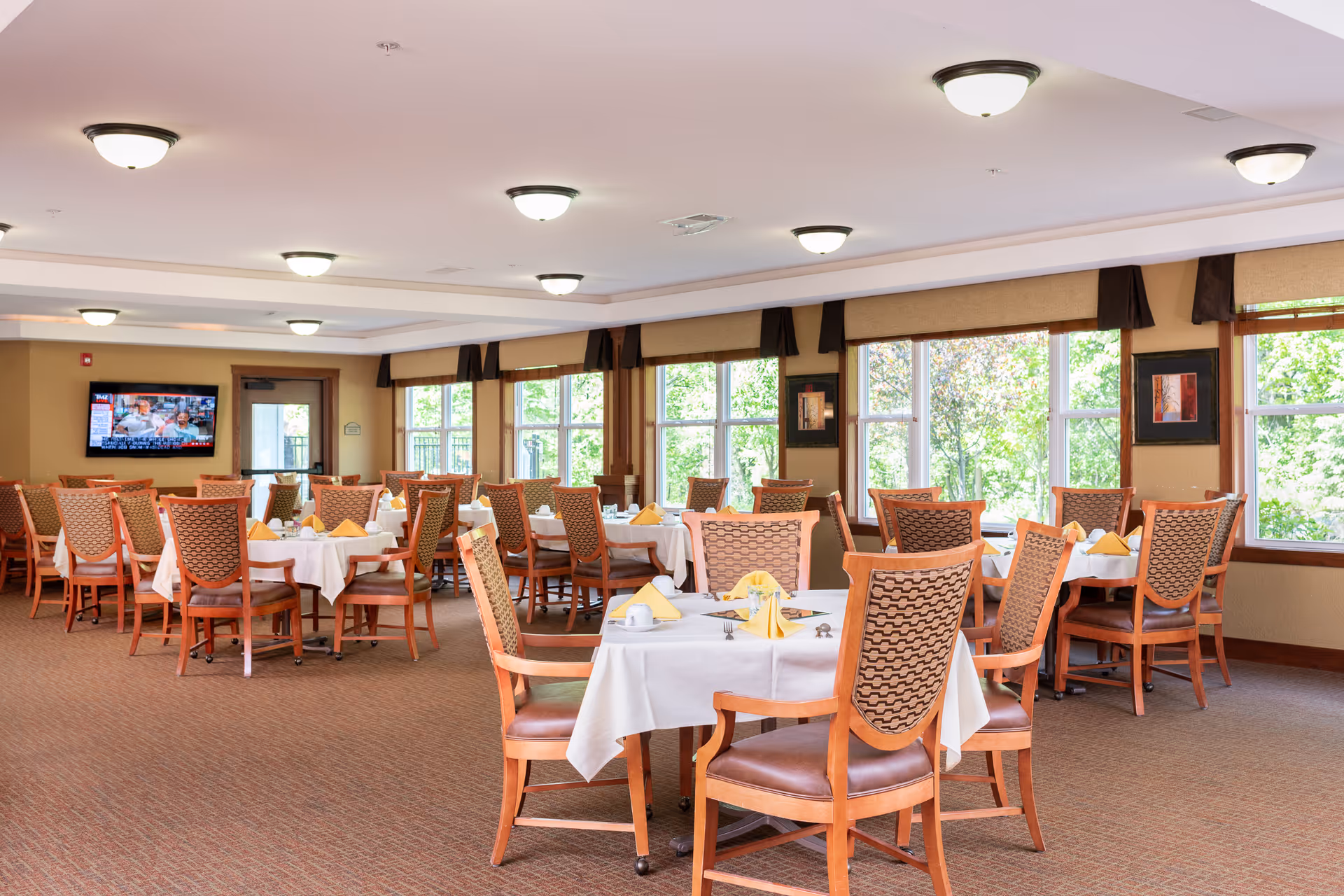 A bright dining room with multiple tables covered in white tablecloths and set with yellow folded napkins, cups, and silverware. The room has large windows letting in natural light and wooden chairs with patterned backs. A television is mounted on the far wall.