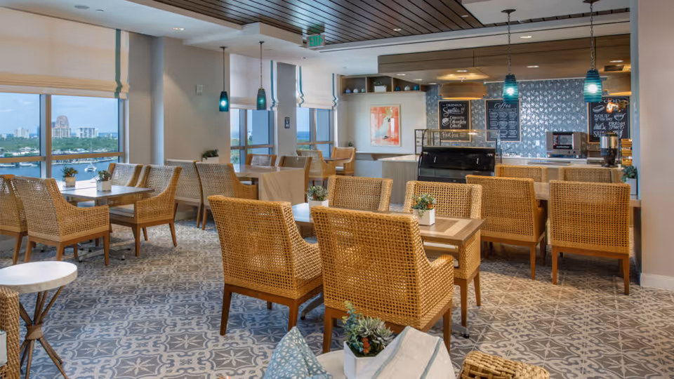 A bright dining area with wicker chairs and wooden tables arranged neatly on a patterned tile floor. Large windows offer a view of a cityscape and water. Pendant lights hang from a wooden ceiling above a counter area with a coffee machine and menu boards on the wall.