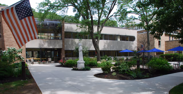 Outdoor courtyard area of a senior living facility with a concrete walkway, landscaped garden beds, a statue in the center, several blue patio umbrellas with tables and chairs, and an American flag on the left side. The building has large windows and is surrounded by trees.