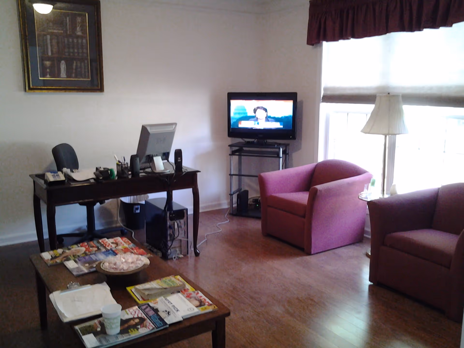 Small living room with hardwood floor featuring two pink armchairs, a TV on a stand, a desk with a computer, and a coffee table covered with magazines.