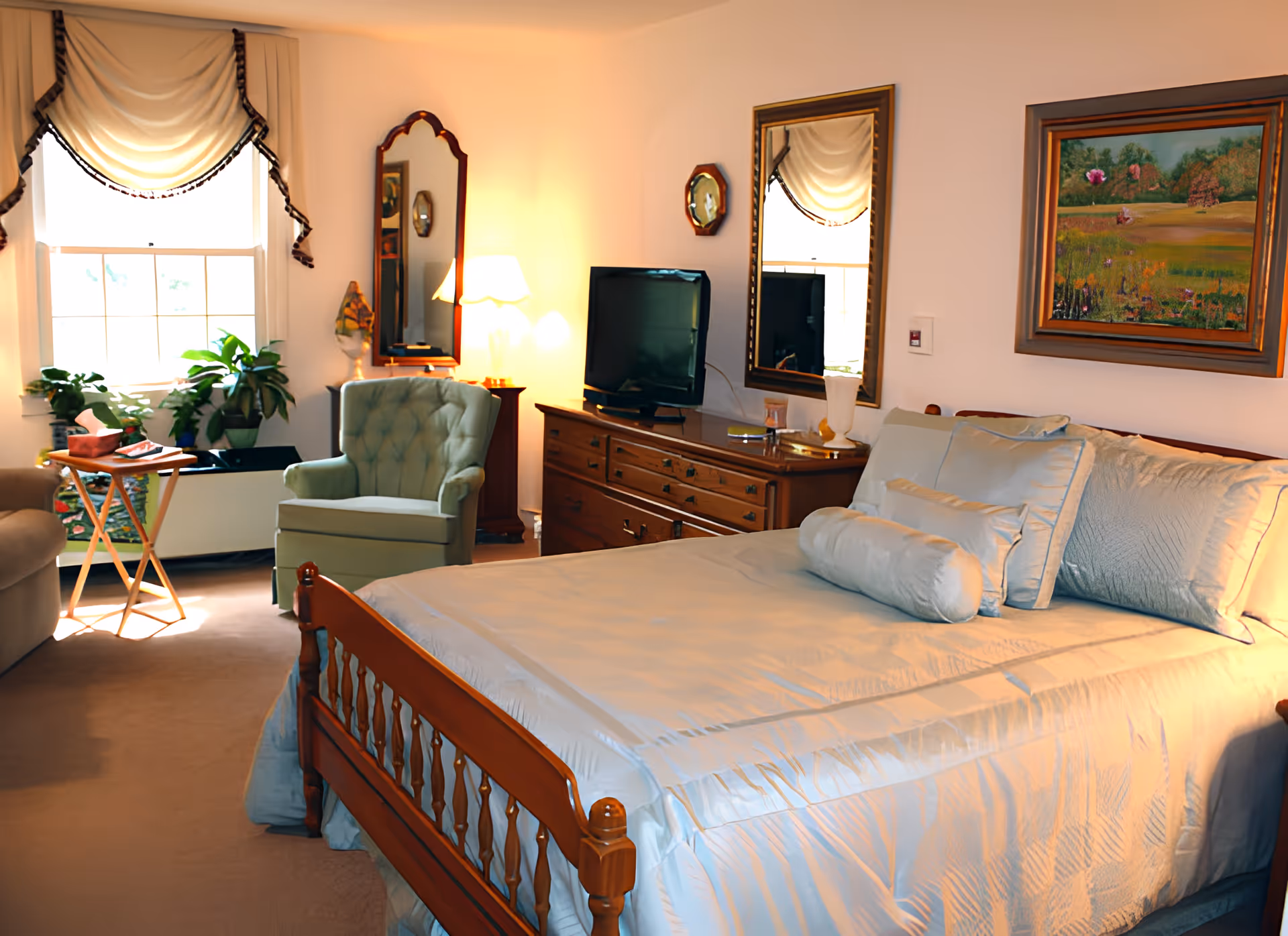 A cozy bedroom in Langdale Place featuring a wooden bed with light blue bedding and multiple pillows. The room includes a wooden dresser with a flat-screen TV on top, two mirrors hanging on the wall, a green upholstered armchair, a small wooden side table, and several potted plants near a window with a decorative valance. A framed painting hangs above the bed, and a lamp provides warm lighting.