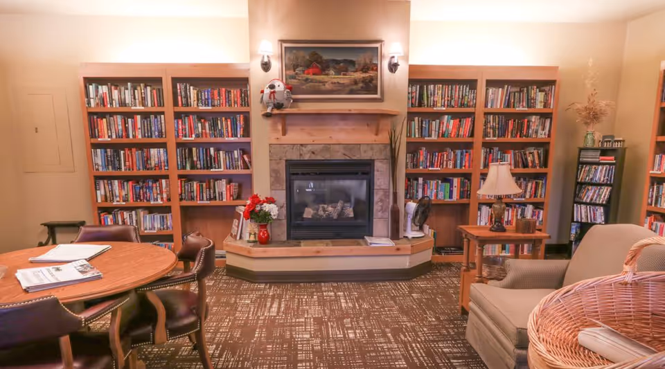 A cozy library room with wooden bookshelves filled with books on either side of a fireplace. The fireplace has a stone surround and a painting above it, flanked by two wall sconces. In front of the fireplace is a small table with a red vase of flowers and some books. To the left, there is a round wooden table with chairs around it and newspapers on top. To the right, there is a comfortable armchair next to a wooden side table with a lamp and a basket in the foreground.