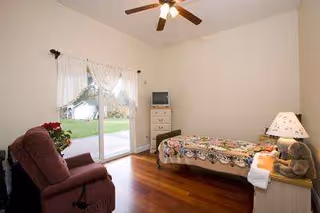 A simple furnished bedroom with a bed, recliner, dresser, ceiling fan, and sliding glass door.
