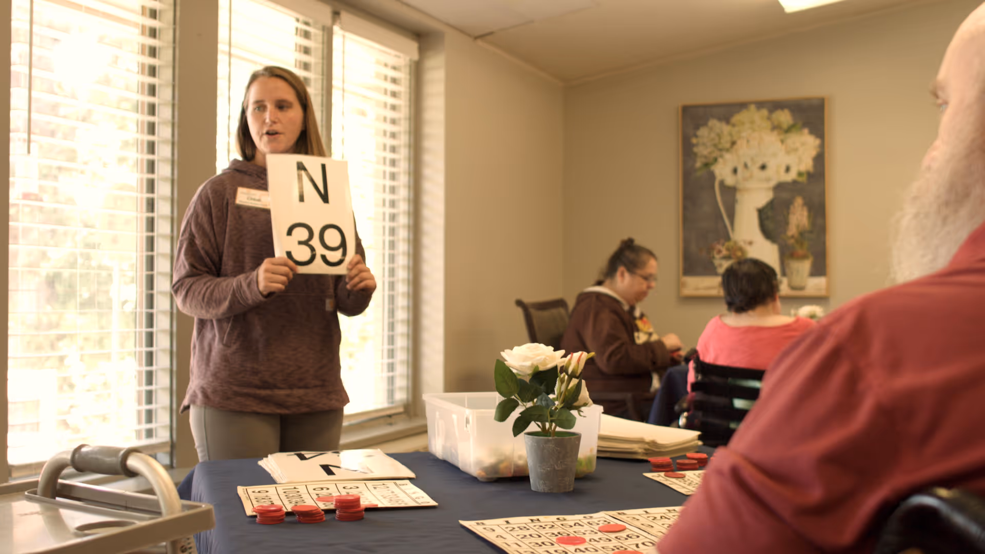 A woman standing and holding a bingo card with the number N39 in a well-lit room with large windows. Several people are seated around a table with bingo cards and red markers, engaged in a bingo game. A potted white rose is on the table, and a floral painting hangs on the wall in the background.