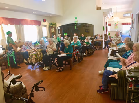 Elderly residents gathered in a communal living room for an activity, many seated in wheelchairs and some wearing green while a musician plays.