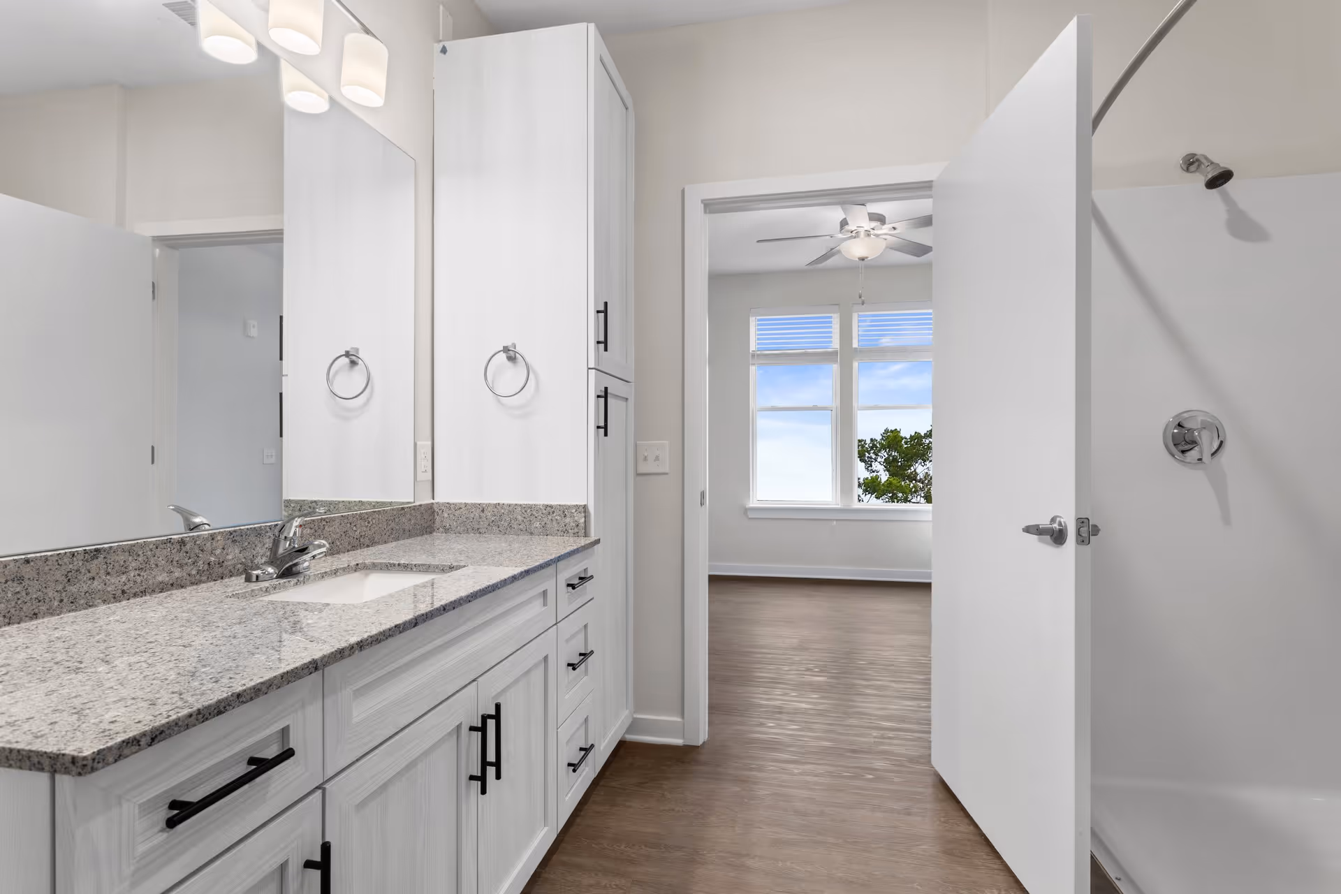 Bright bathroom with a granite-topped vanity, large mirror, and an open doorway leading to a bedroom.