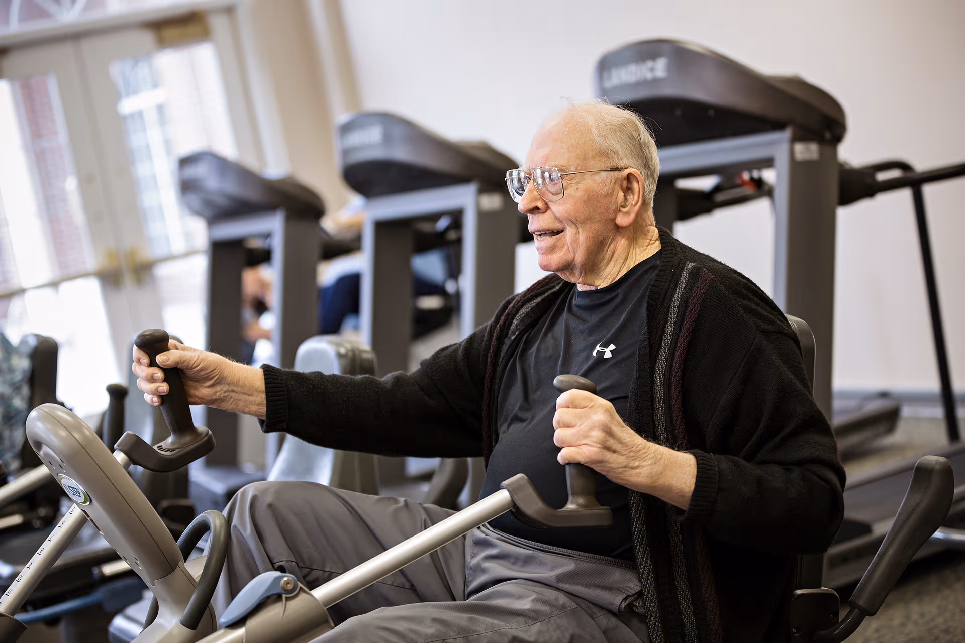 An elderly man wearing glasses and a black sweater is exercising on a seated elliptical machine in a fitness room with treadmills in the background.