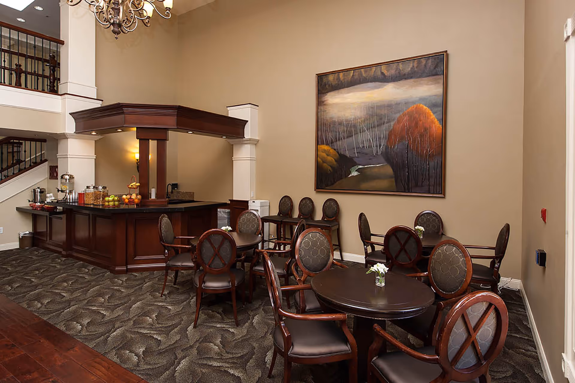 Communal dining/lounge area with round tables and chairs and a dark wood service bar under a large landscape painting.