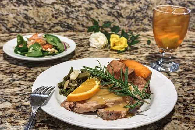 A plated meal on a granite countertop featuring sliced turkey with gravy, a sprig of rosemary, a slice of orange, green beans with almond slices, and a baked sweet potato. In the background, there is a small salad on a white plate and a glass of iced tea with lemon. Decorative flowers are placed behind the plates.