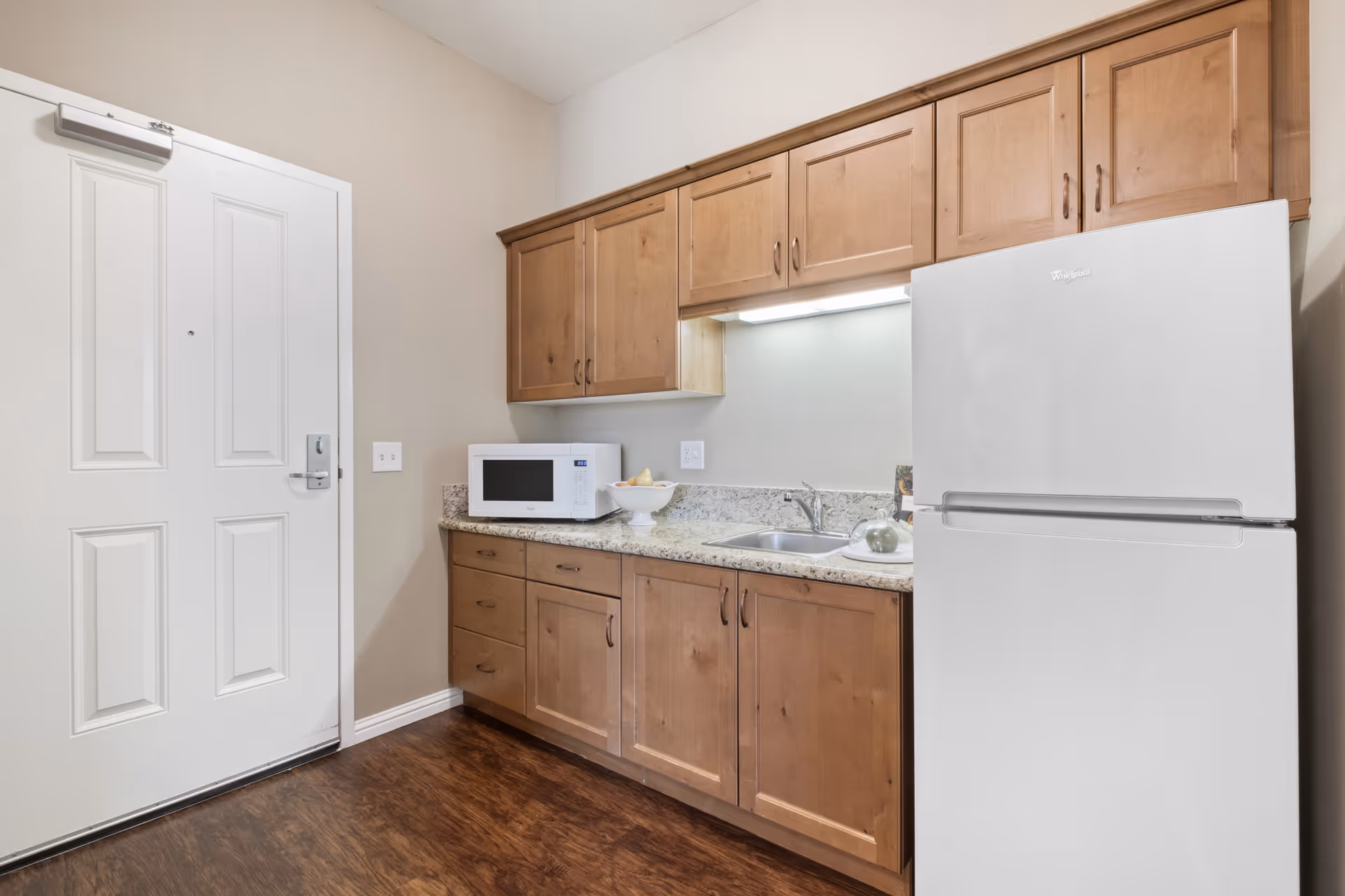 A small kitchen area with wooden cabinets, a white refrigerator, a microwave on the countertop, a sink, and a bowl of fruit. The floor is wooden and there is a white door to the left.