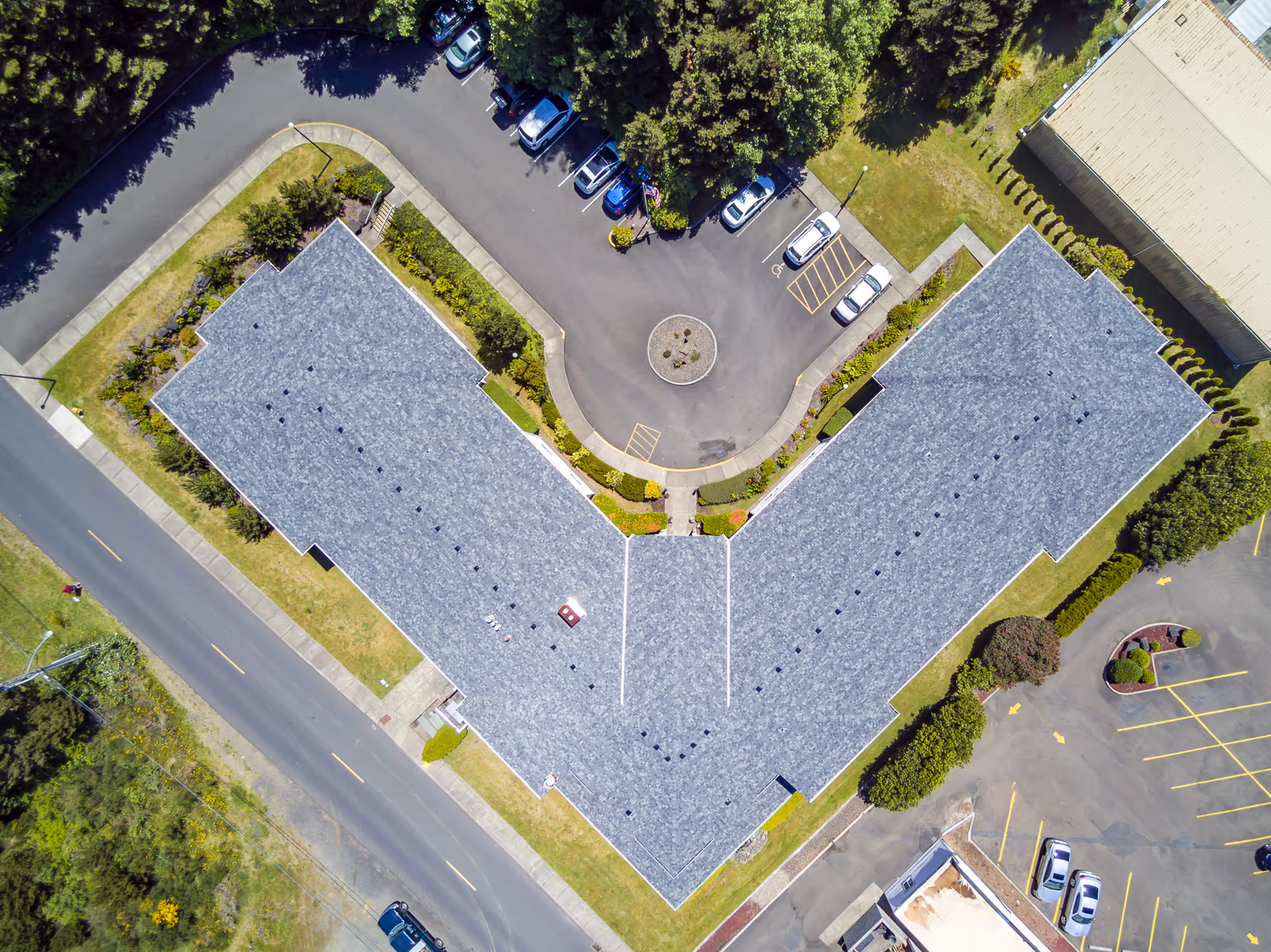 Aerial view of a V-shaped building with a gray roof surrounded by parking lots, roads, and greenery. Several cars are parked in the parking areas adjacent to the building.