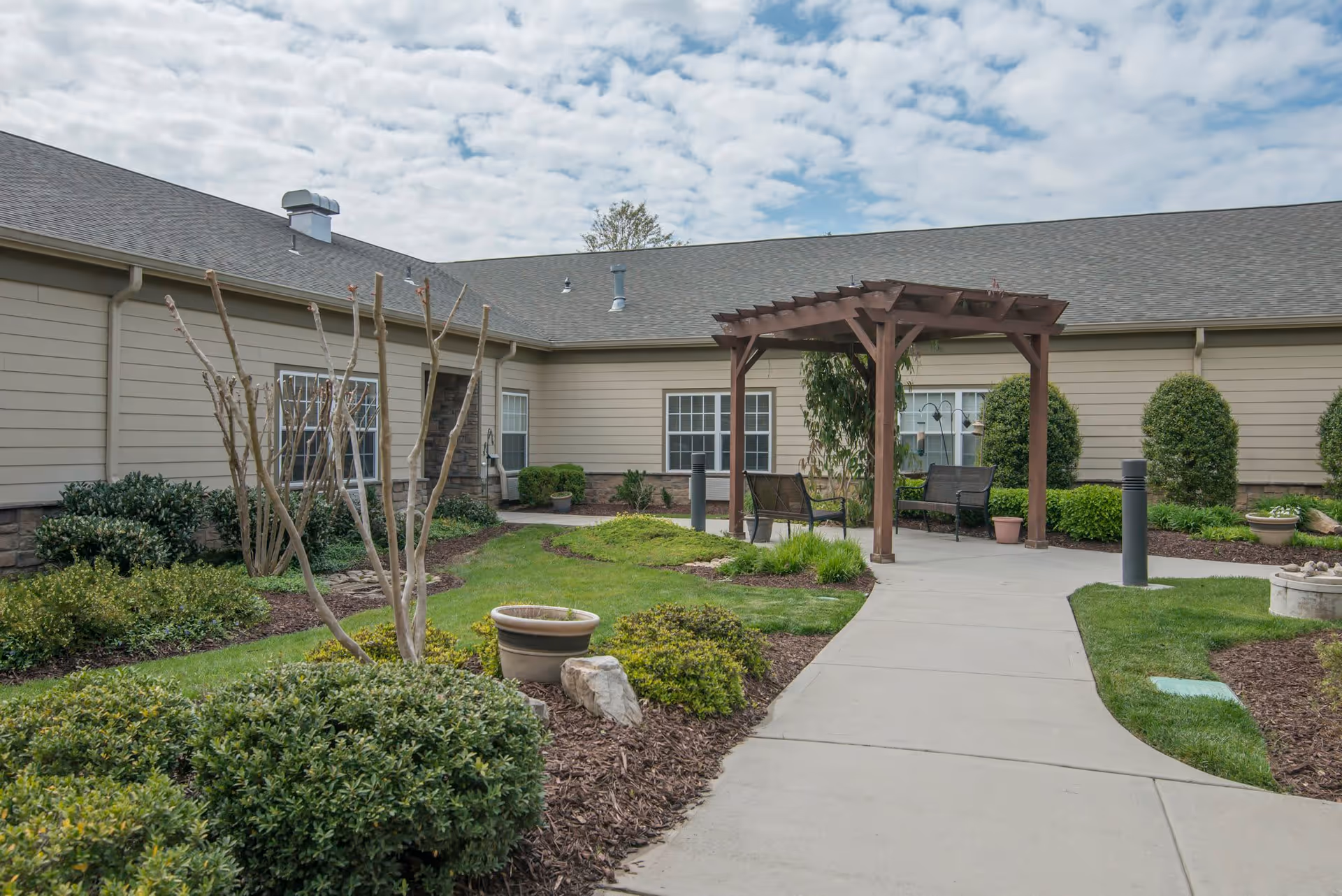 Outdoor courtyard area at Morning Pointe Of Greenbriar Cove featuring a paved walkway, green grass, trimmed bushes, a wooden pergola with seating underneath, and beige building walls with windows in the background under a partly cloudy sky.