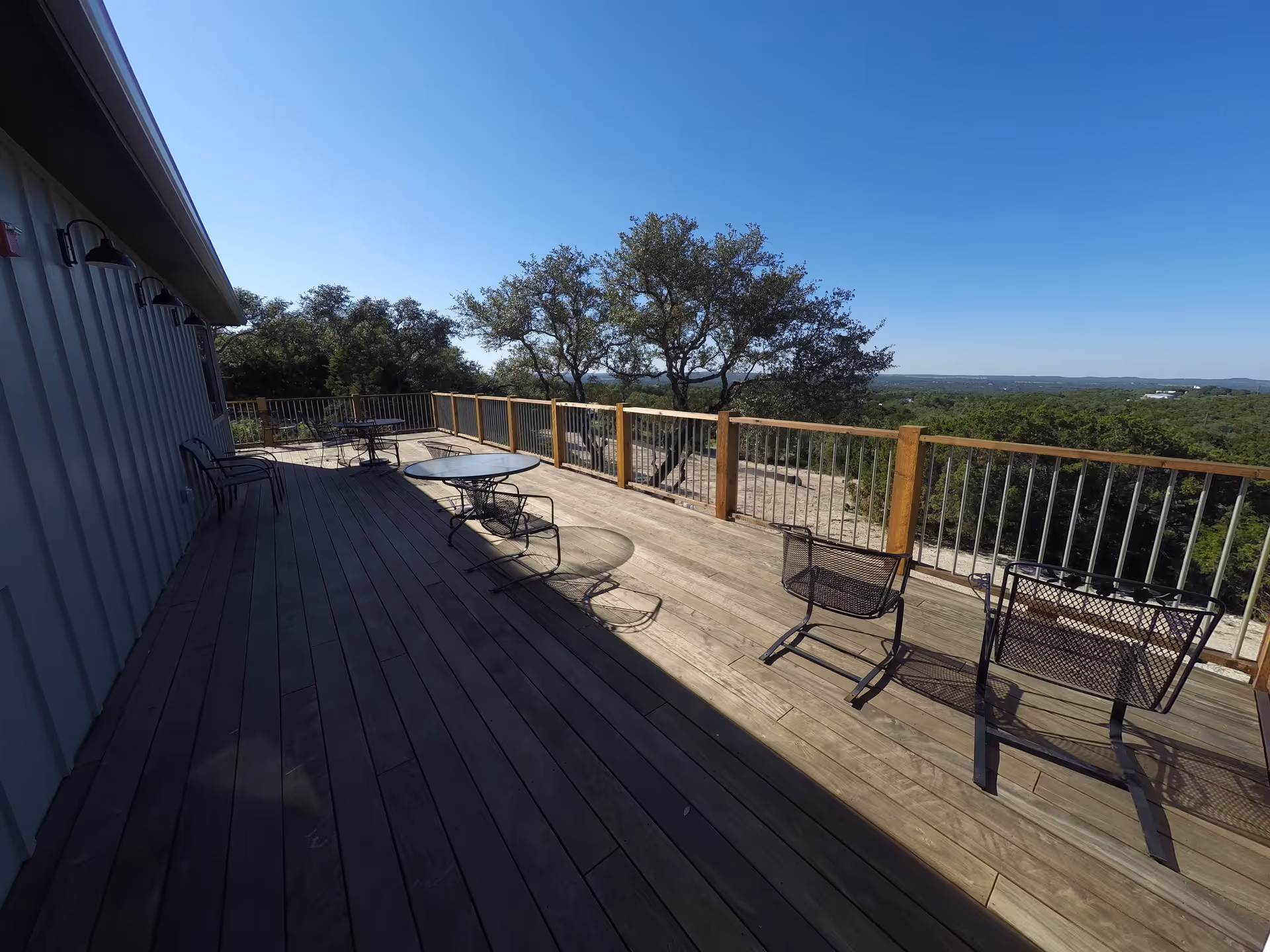 Outdoor wooden deck with metal chairs and tables, overlooking a scenic view of trees and hills under a clear blue sky.
