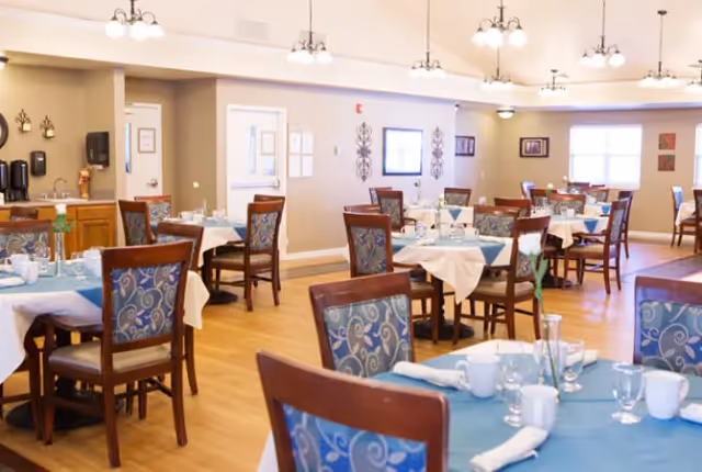 A bright and spacious dining room with multiple tables covered in white and blue tablecloths, set with cups, glasses, and napkins. Wooden chairs with blue patterned cushions surround the tables. The room has wooden flooring, beige walls, and several light fixtures hanging from the ceiling. Wall decorations and windows provide natural light.