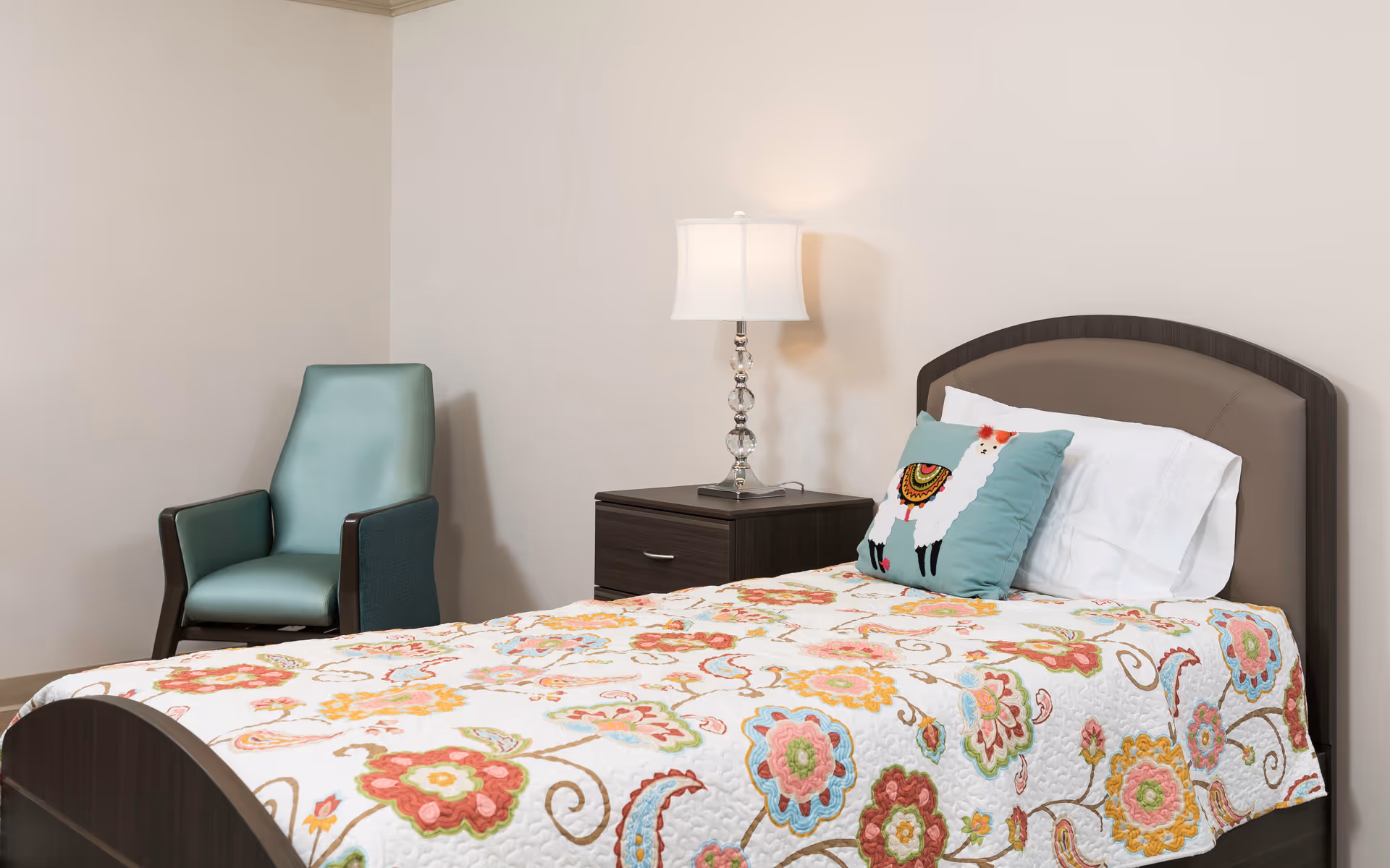 A neatly made single bed with a colorful floral quilt and a decorative pillow featuring a llama design. Next to the bed is a dark wooden nightstand with a clear glass lamp on top. A teal upholstered armchair is positioned against the beige wall in the corner of the room.