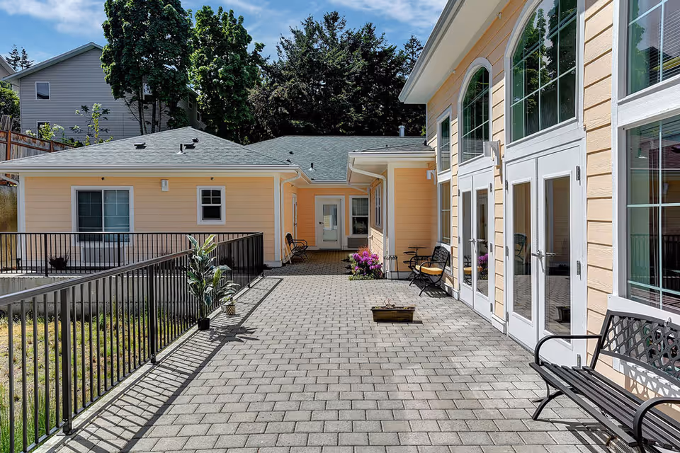 Outdoor patio area at Tabor Crest Memory Care with paved stone flooring, black metal benches, potted plants, and light yellow building walls with large windows and doors.