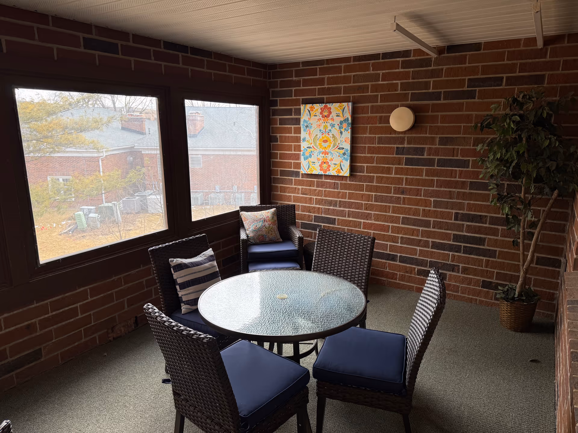 Enclosed brick sunroom with a round glass-top table and four wicker chairs with blue cushions, wall art, and a potted plant.