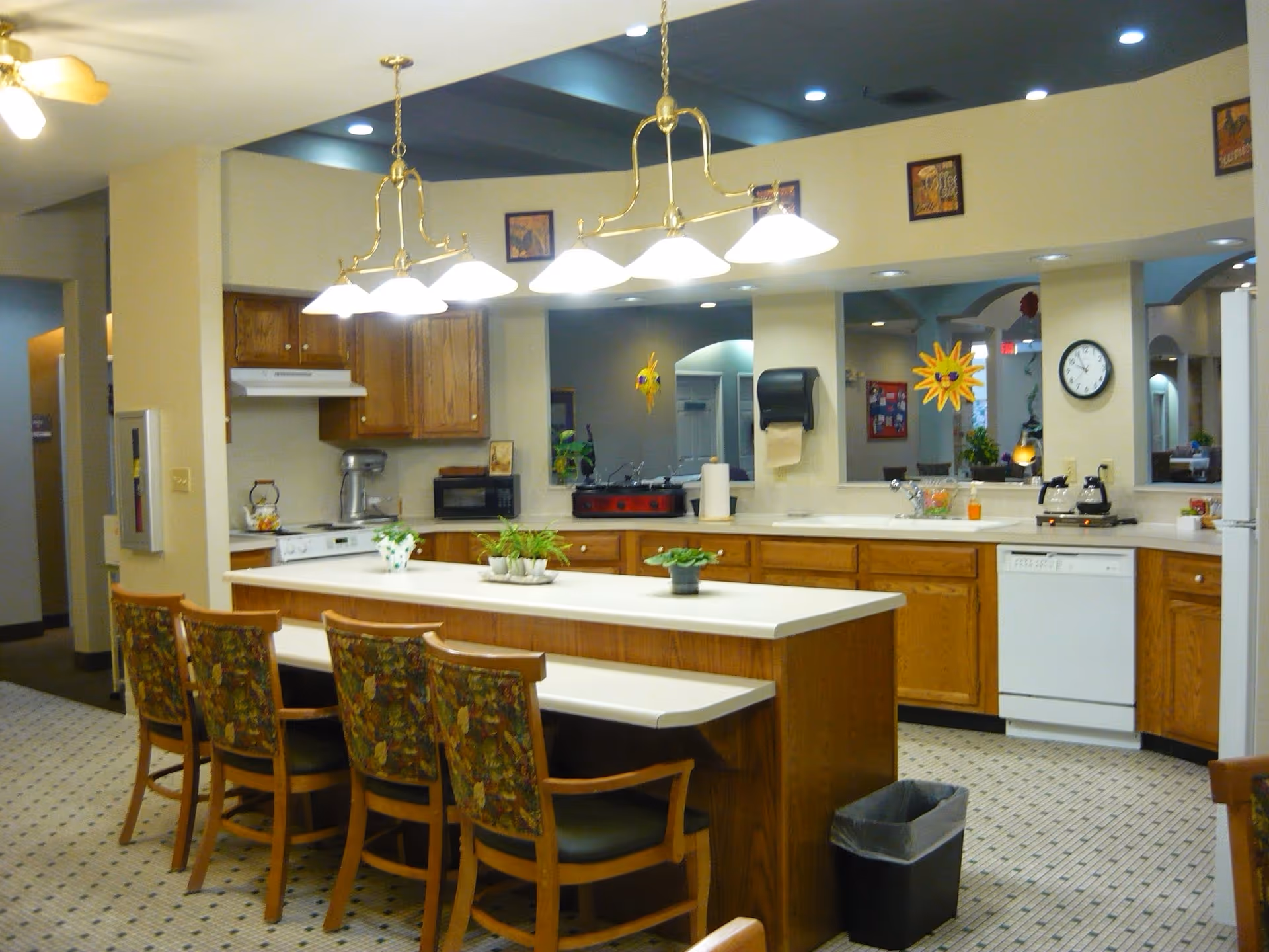 Bright communal kitchen and dining area with a central island, pendant lights, wooden cabinets, and several chairs.
