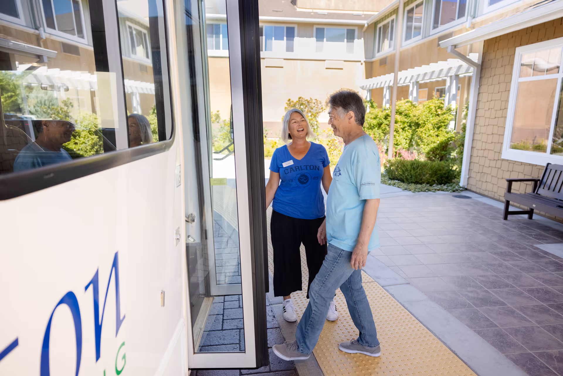 An elderly man and a woman wearing a Carlton Senior Living shirt are standing outside near a building entrance and a shuttle bus, smiling and engaging in conversation on a sunny day.