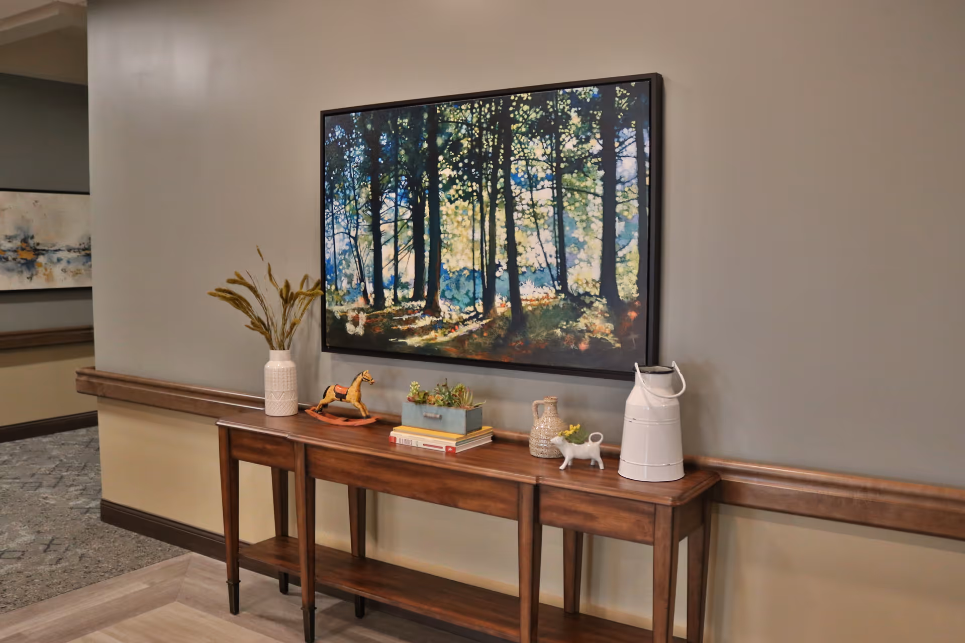 A hallway console table with decorative items beneath a large framed forest painting on a muted gray wall.
