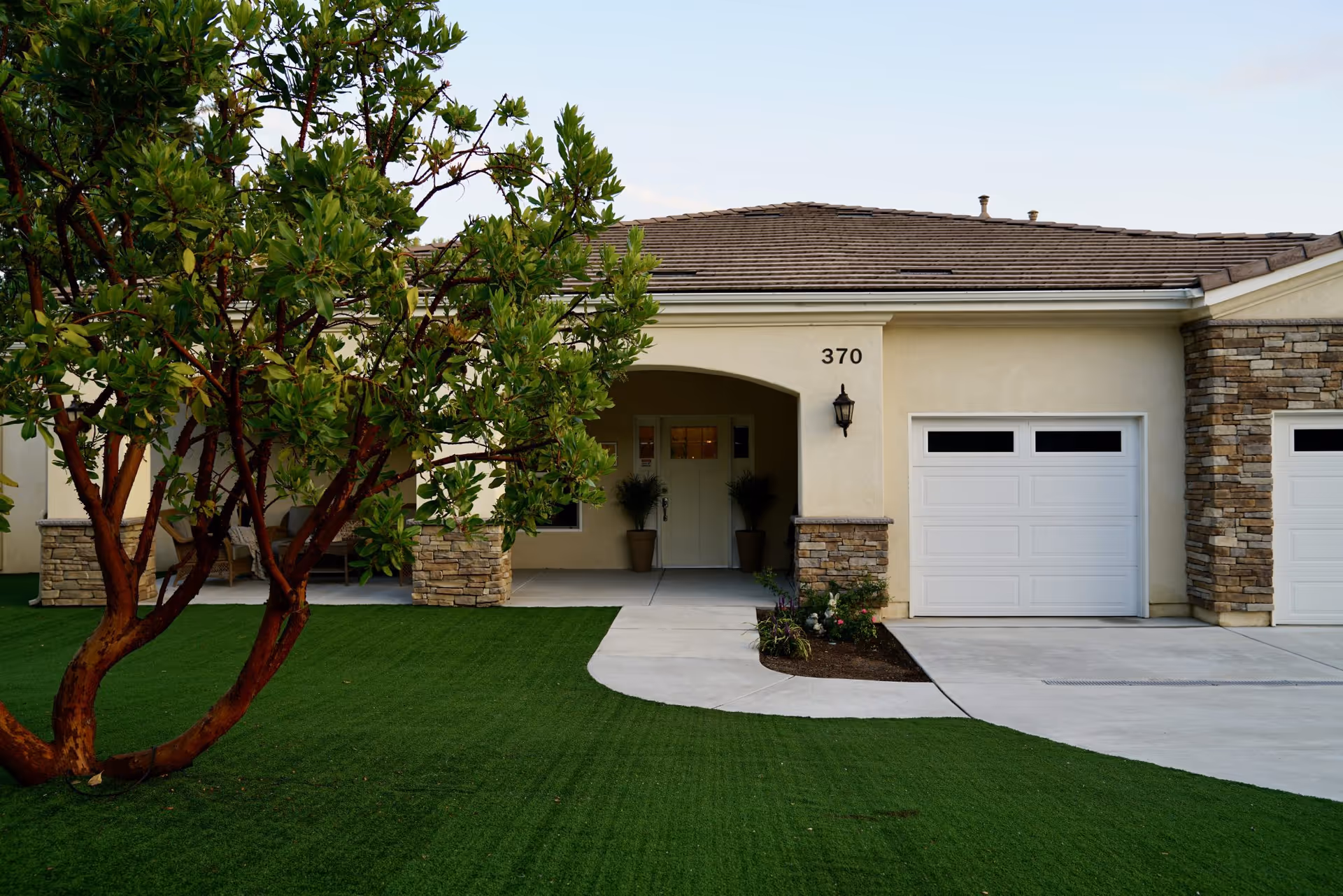 Front exterior view of a single-story senior living facility building with beige walls, a tiled roof, a white garage door, and a covered entrance with two potted plants. The building number 370 is visible above the entrance. There is a green lawn and a tree in the foreground.