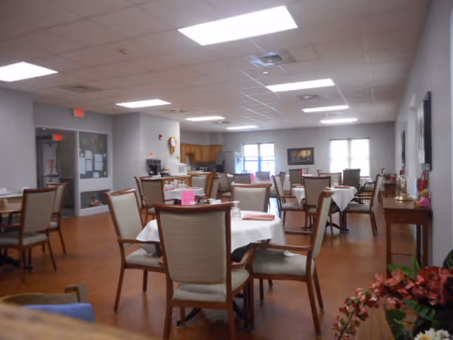A dining room in an assisted living facility with multiple round tables covered with white tablecloths and surrounded by beige cushioned chairs. The room has wooden flooring, fluorescent ceiling lights, and a small kitchenette area in the background. There are windows letting in natural light and some framed pictures on the walls.