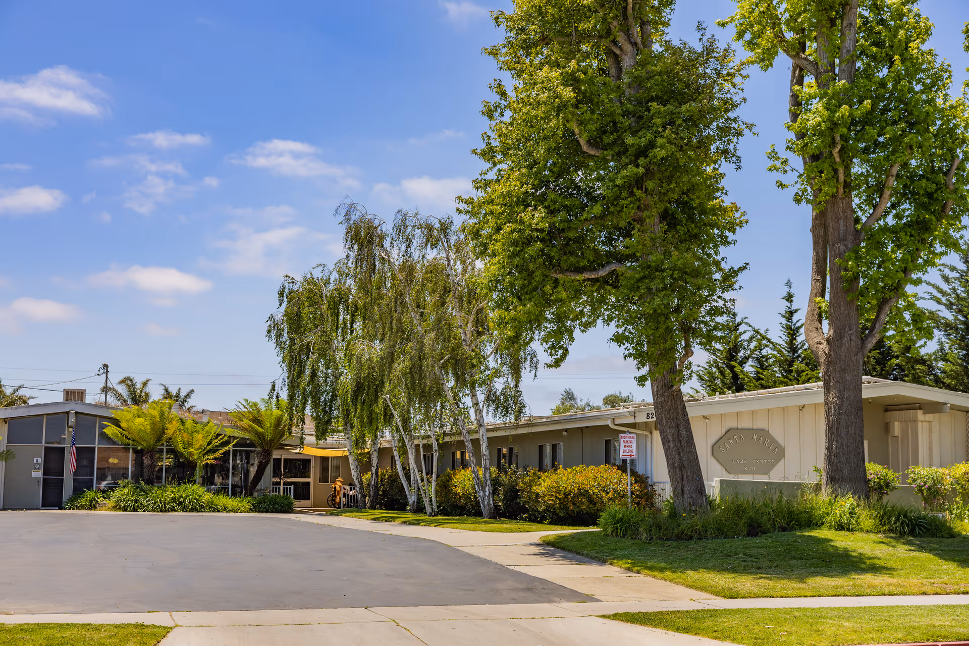 Front view of the Santa Maria Post Acute single-story building with trees, lawn, and a driveway under a blue sky.