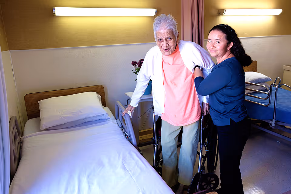 An elderly woman is being assisted by a caregiver in a room with two hospital beds. The elderly woman is standing with the help of a walker, while the caregiver supports her from behind. The room has soft lighting and a small table with flowers between the beds.