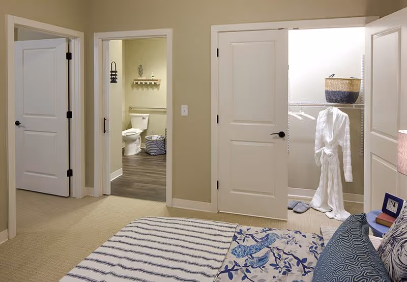 View of a bedroom with a bed covered in a blue and white patterned blanket and pillows. The room has beige walls and carpet. There are three doors visible: one leading to a bathroom with a toilet and shelf, one to a closet with a white robe hanging and a basket on a shelf, and one closed door.