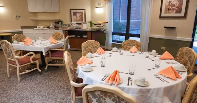 A dining room with two tables set for a meal. One table is round and the other is rectangular, both covered with white tablecloths and set with plates, cups, glasses, silverware, and folded peach-colored napkins. The room has floral upholstered chairs, a carpeted floor, framed artwork on the walls, and large windows with curtains letting in natural light. A buffet area with food and beverages is visible in the background.