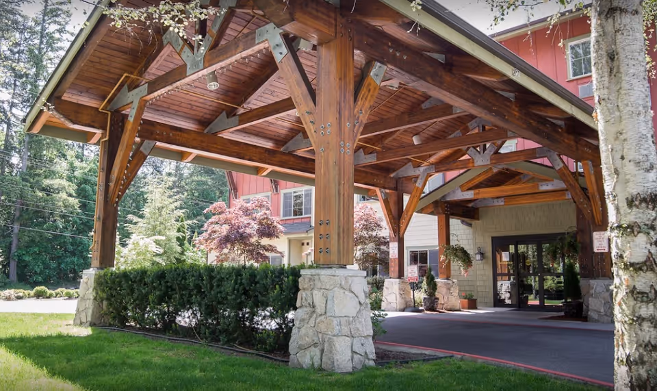 Covered entrance of a senior living facility with wooden beams and stone pillars, surrounded by greenery and trees, leading to glass doors of the building.