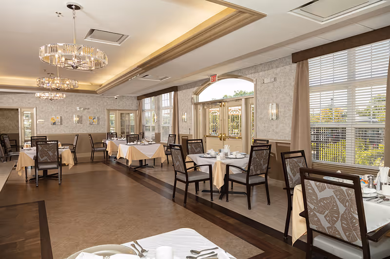 A bright and spacious dining room in an assisted living facility with multiple tables covered with white and beige tablecloths, set with plates, cups, and utensils. The room features large windows with blinds and curtains, elegant chandeliers, and double glass doors leading outside.