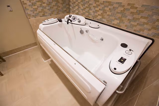 A white walk-in bathtub with built-in controls and safety handles, installed in a bathroom with beige tiled walls and floor.