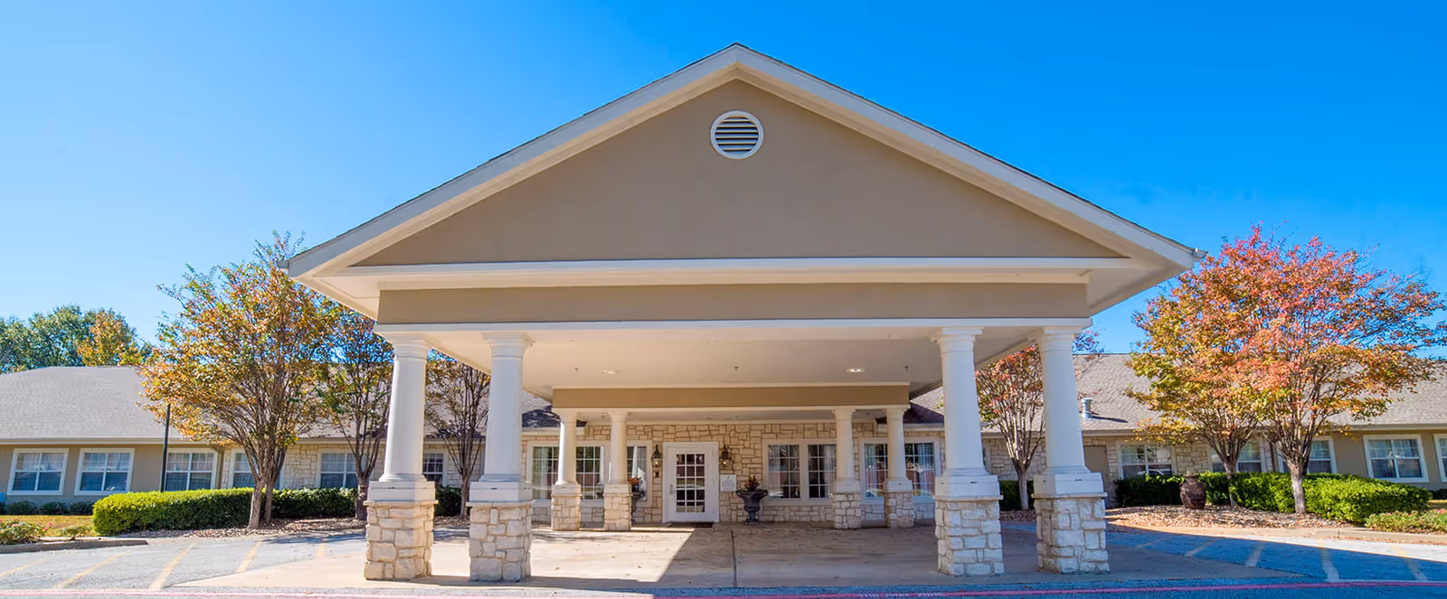 Front exterior view of Brookdale Tyler South facility with a covered entrance supported by white columns and stone bases, surrounded by trees with autumn foliage under a clear blue sky.